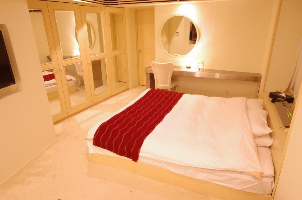 A hotel room with a bed, a round mirror, a desk, and a wardrobe with glass doors.