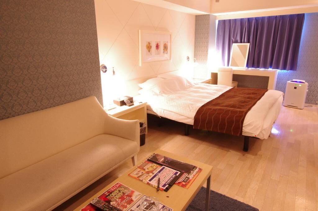 A hotel room with a bed, a sofa, a bedside table, a lamp, a mirror, and a small table with magazines.