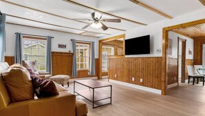 A spacious living room with a sofa, a coffee table, a ceiling fan, wooden walls, a TV mounted on the wall, and large windows. - Charming Home Near Stockyards & Trinity Trails Central Fort Worth (River Oaks)