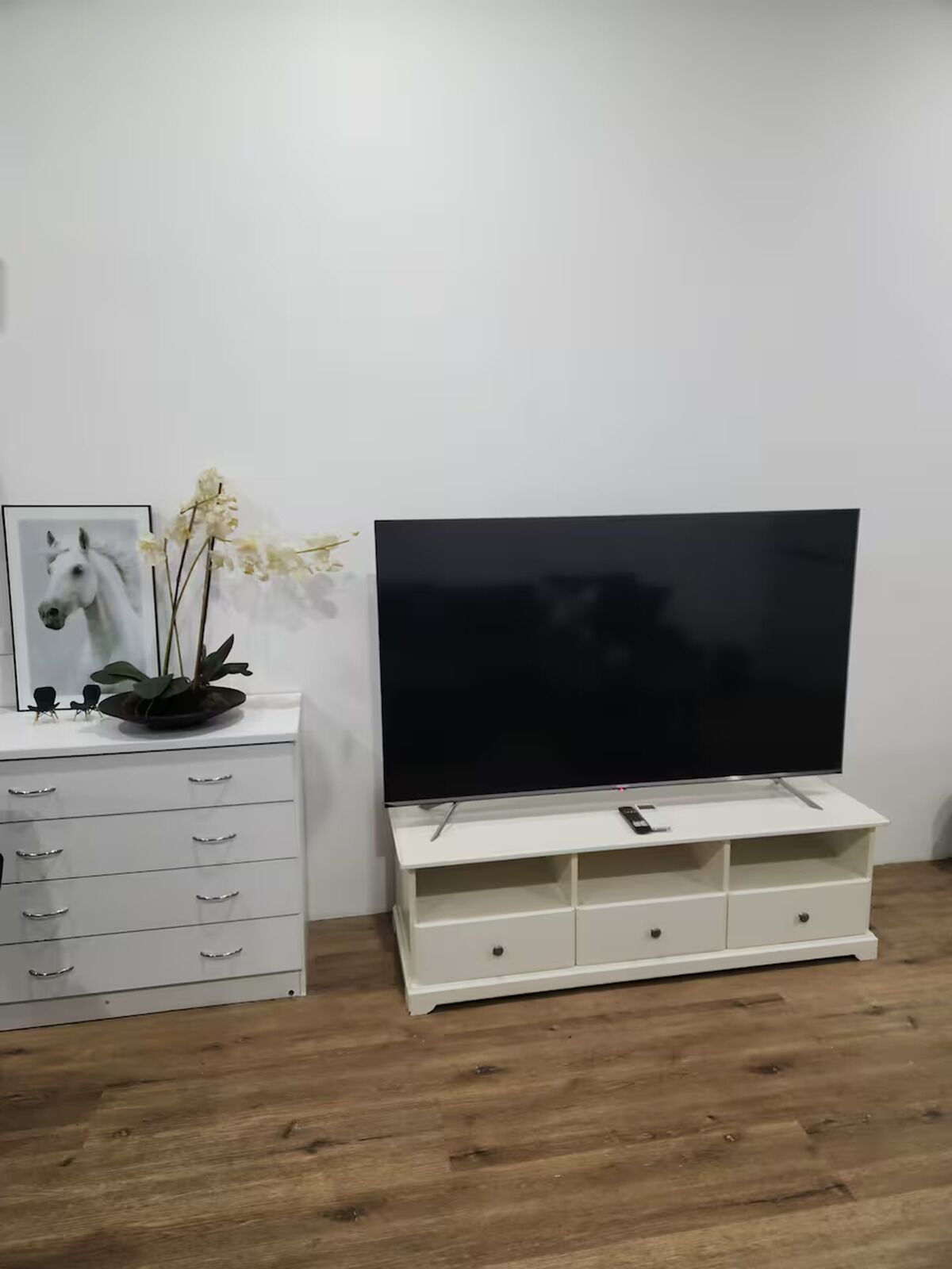 Un mueble de entretenimiento blanco con un televisor de pantalla plana, un cuadro de un caballo enmarco y una orquídea en maceta.