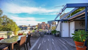 A rooftop terrace with a glass table, wicker chairs, potted plants, and a view of city buildings. - New York-Style Loft in Sydney CBD (Darlinghurst)