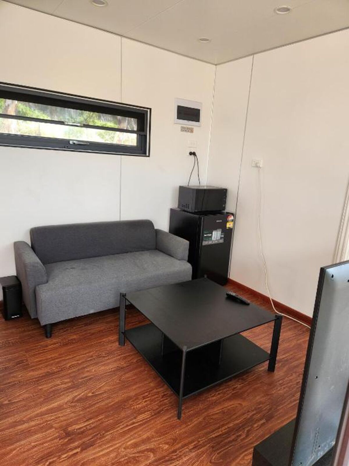 Una habitación con un sofá gris, una mesa de centro negra, un televisor sobre un mueble bajo y una ventana con vistas a zonas verdes.