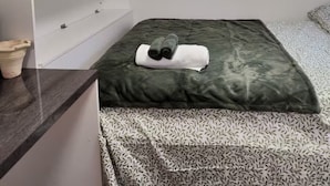 Una habitación con una cama, un televisor de pantalla plana sobre una estantería blanca, un equipo de aire acondicionado y una mesita con un objeto decorativo.