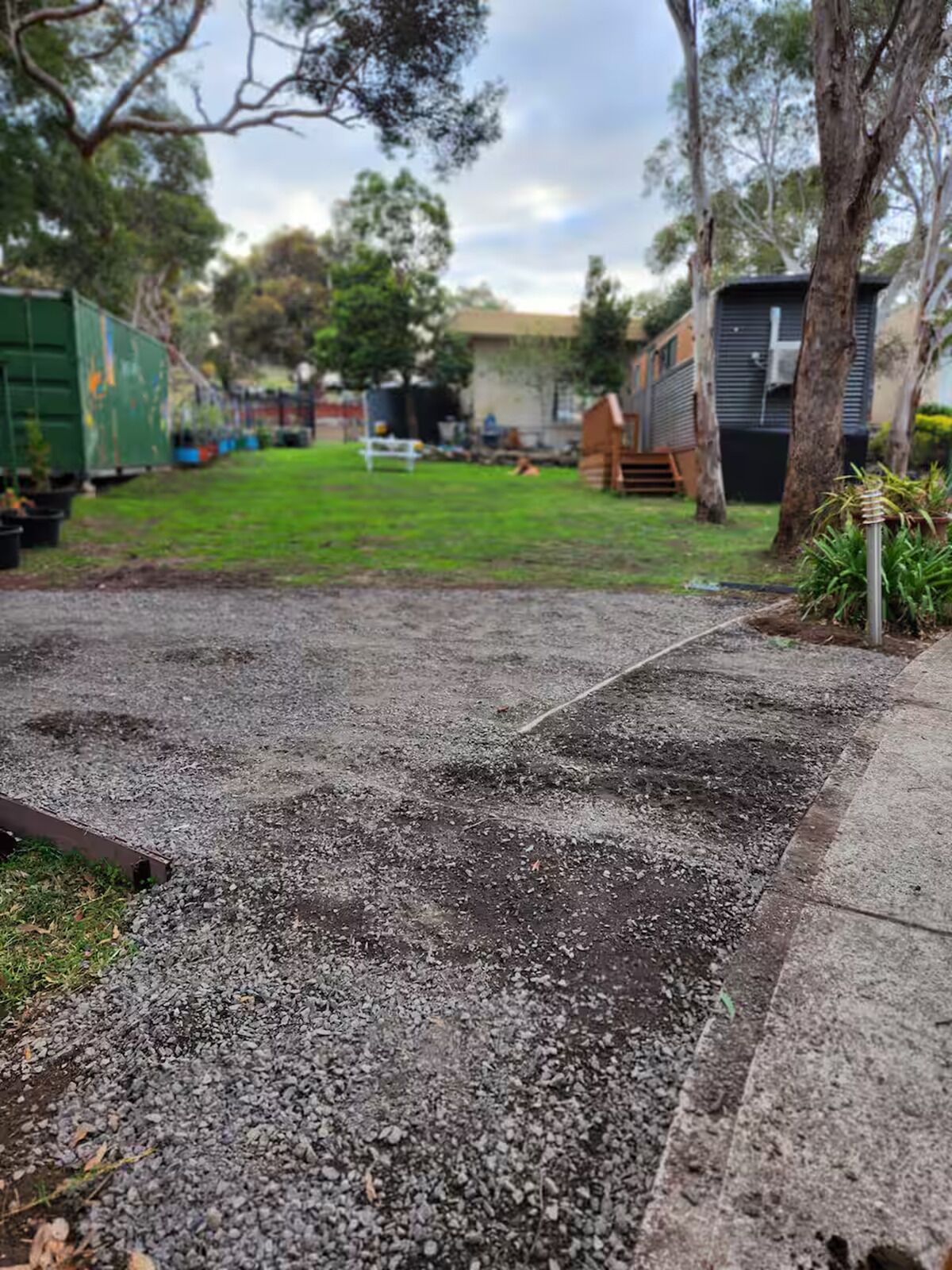 A gravel driveway leading to a residential area with a green truck parked on the left, a small wooden structure on the right, and a grassy yard in the background.
