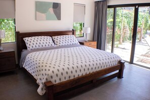 A bedroom with a large bed, a nightstand, a lamp, a window with curtains, and a view of greenery outside. - Golden Shore Villa in Tamarindo (Tamarindo)