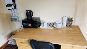 A wooden desk with a black chair, a coffee machine, a kettle, a tray with cups and a plate, and a set of cutlery.