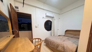 Une petite pièce comprenant une table en bois, un lit simple, une chaise, une fenêtre ronde et une porte.