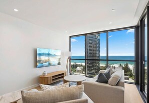 A modern living room with a large window, a flat-screen TV, a round coffee table, and a sofa with cushions. - Yves 2 Bedroom Ocean (Mermaid Beach)