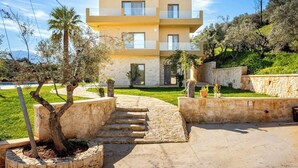 A modern two-story building with a stone facade, a central entrance with steps, and a surrounding landscaped garden with trees and shrubs. - Beautiful apartment for the whole family with view at the beach (Stalos)