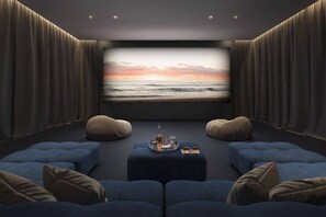 Une pièce sombre, avec un grand écran diffusant une image de coucher de soleil sur l’océan, des sièges bleus et un plateau contenant des verres et une bouteille.