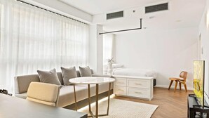 Una sala moderna con un sofá, una mesa de centro redonda, un aparador blanco, un televisor y un ventanal con cortinas translúcidas.