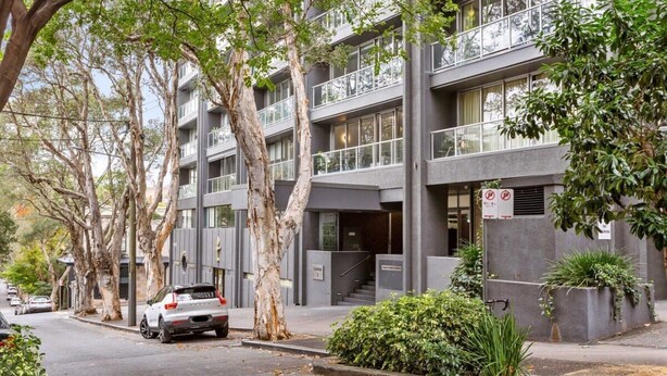 Un edificio de varios pisos con balcones, un coche estacionado adelante y árboles a lo largo de la calle.