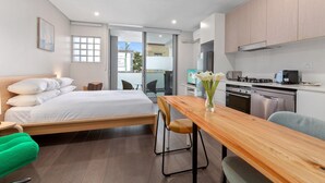 A modern hotel room with a bed, a wooden dining table, a kitchen area, and a balcony with a view.