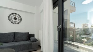 Un soggiorno moderno con un divano grigio, un orologio rotondo a parete e vista sulla città attraverso una porta in vetro.