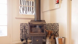 A wood-burning stove with a metal basket and bucket beside it, a patterned wall, and a window with blinds. - Towncottage Tenterfield Stay with Scenic Views (Tenterfield)