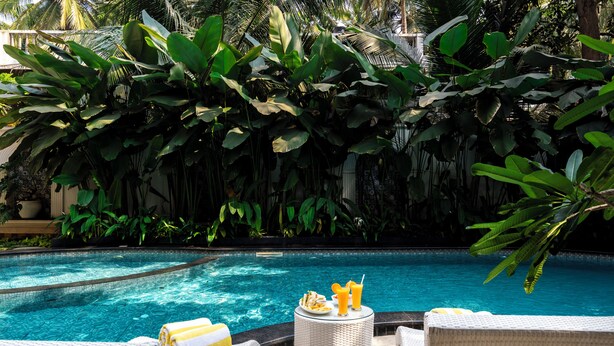 A poolside area with lounge chairs, a table with drinks, and lush tropical foliage. - Staymaster Veridian 2BR - Standard (Calangute)