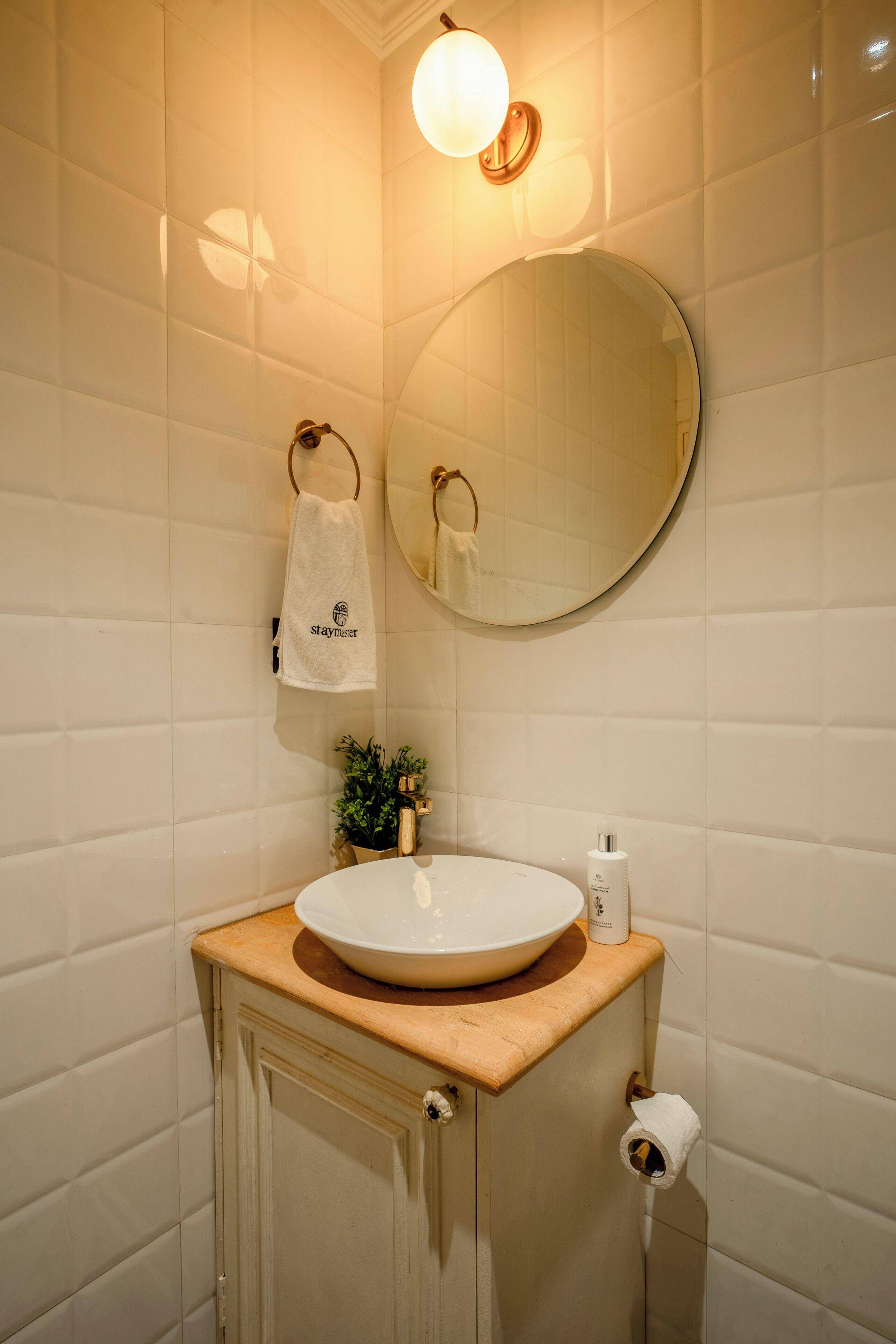 Ein Badezimmer mit rundem Spiegel, Waschbecken mit Holzablage, Handtuch auf einem Handtuchhalter und Toilettenpapierhalter.