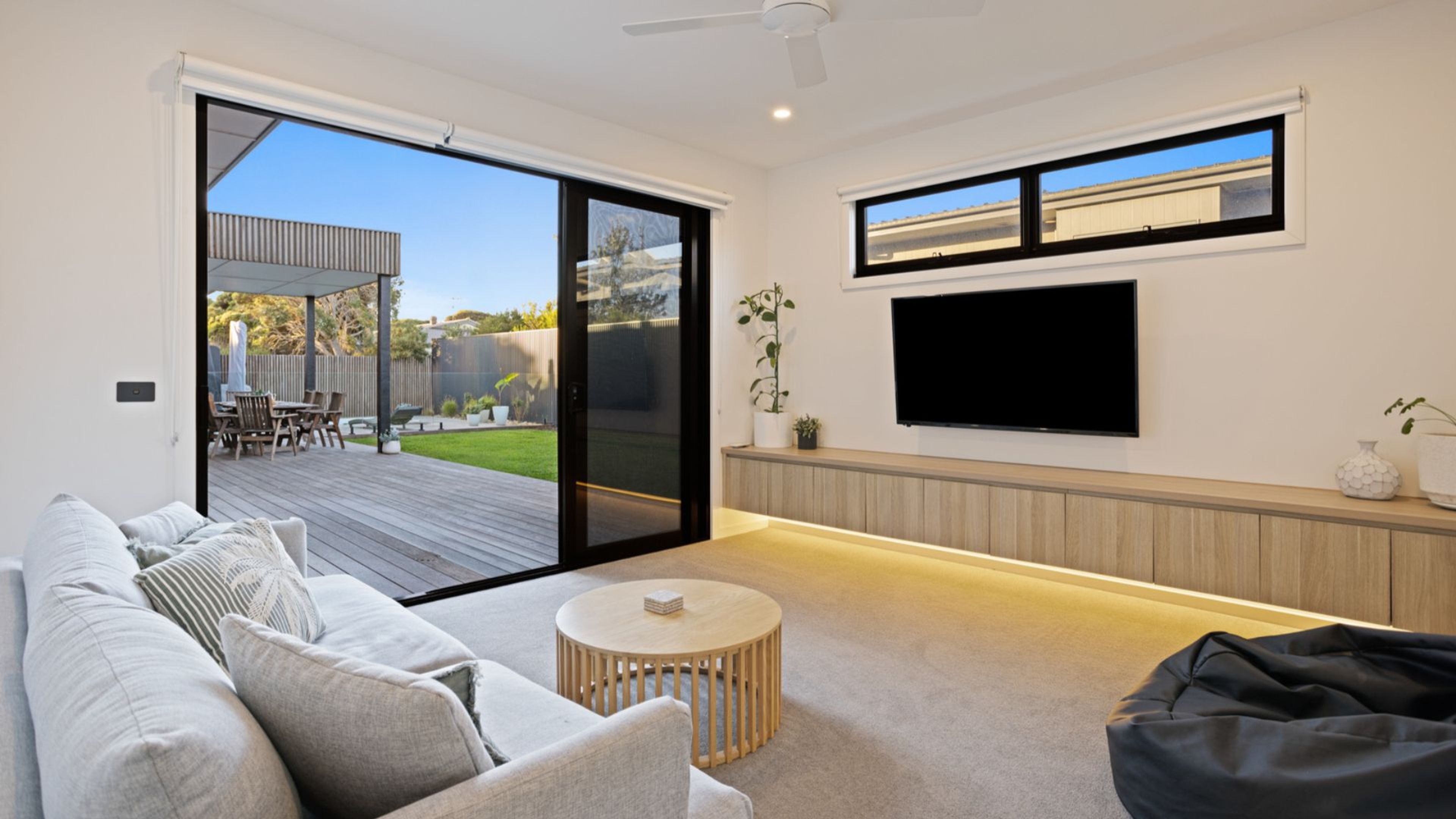 A modern living room with a sofa, a round coffee table, a flat-screen TV mounted on the wall, and a sliding glass door leading to an outdoor patio.
