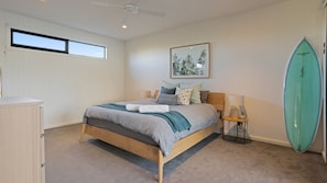 A bedroom with a bed, a ceiling fan, a window, a framed picture, a surfboard, and a small table with a lamp. - Salty Sixteen Rye Retreat with Pool (Rye)