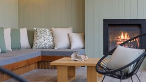 A cozy seating area with a sofa, cushions, a table, and a chair. A fireplace with a visible fire is present. - Salty Sixteen Rye Retreat with Pool (Rye)