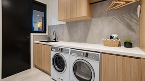 A modern laundry room with two front-loading washing machines, a sink, and wooden cabinets. - Salty Sixteen Rye Retreat with Pool (Rye)