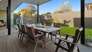 An outdoor dining area with a wooden table and chairs, surrounded by a wooden deck and a fenced backyard. - Salty Sixteen Rye Retreat with Pool (Rye)