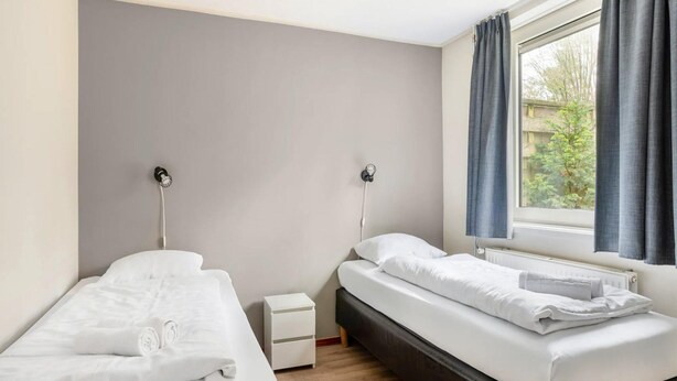 A bedroom with two beds, a nightstand, a window with curtains, and a view of greenery outside. - 6-person Comfort Bungalow With Sauna (Kootwijk)