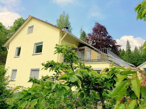 Ein zweistöckiges Gebäude mit Balkon und einer sichtbaren Treppe, davor ein Baum.