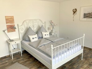 Ein ordentlich eingerichtetes Schlafzimmer mit weißem Metallbettgestell, grauem Bettzeug und einem Nachttisch mit Lampe.