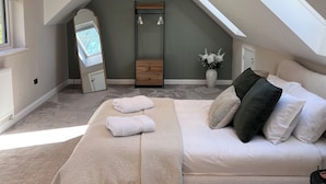 Un dormitorio con techo inclinado, una cama con cubrecama color beige, una mesita de noche, un espejo y una ventana.