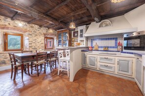 Une cuisine rustique avec un mur en pierre, des poutres apparentes, une table à manger avec des chaises et un crédence carrelée blanche et bleue intégrée.