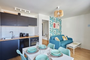 Eine kompakte Küche mit modernen Geräten, ein Essbereich mit türkisfarbenem Geschirr und ein Wohnbereich mit blauem Sofa und gemustertem Wandpapier.