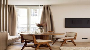 Ein moderner Essbereich mit einem runden Tisch, zwei Holzstühlen, einer Blumenvase und Blick durch große Fenster nach draußen.