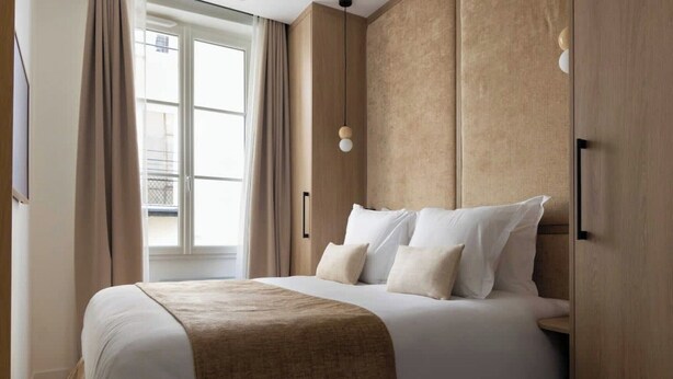 Ein ordentlich bezogenes Bett mit beiger Bettdecke und Kissen, ein hölzernes Kopfteil und ein Fenster mit Vorhängen.