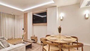 Un salon moderne comprenant un coin repas, un téléviseur à écran plat fixé au mur et une petite table d’appoint sur laquelle est posé un vase de fleurs.