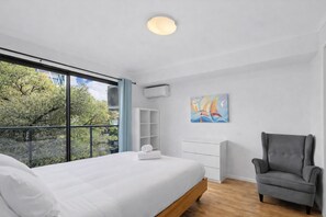 A modern bedroom with a large window, a bed, a chair, a wall-mounted air conditioner, and a colorful painting. - Stylish Bowen Hills Studio Near City Centre (Bowen Hills)