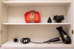 Une étagère sur laquelle se trouvent une trousse de premiers secours rouge, un sèche-cheveux noir et un objet noir doté de plusieurs pointes.