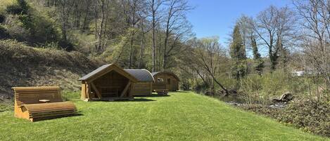 A wooden cabin with a sloped roof, a bench, and a small table in a grassy area with trees in the background.