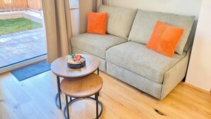 Un sofá gris con cojines naranjas, una pequeña mesa redonda de madera con un frutero, y un suelo de madera.