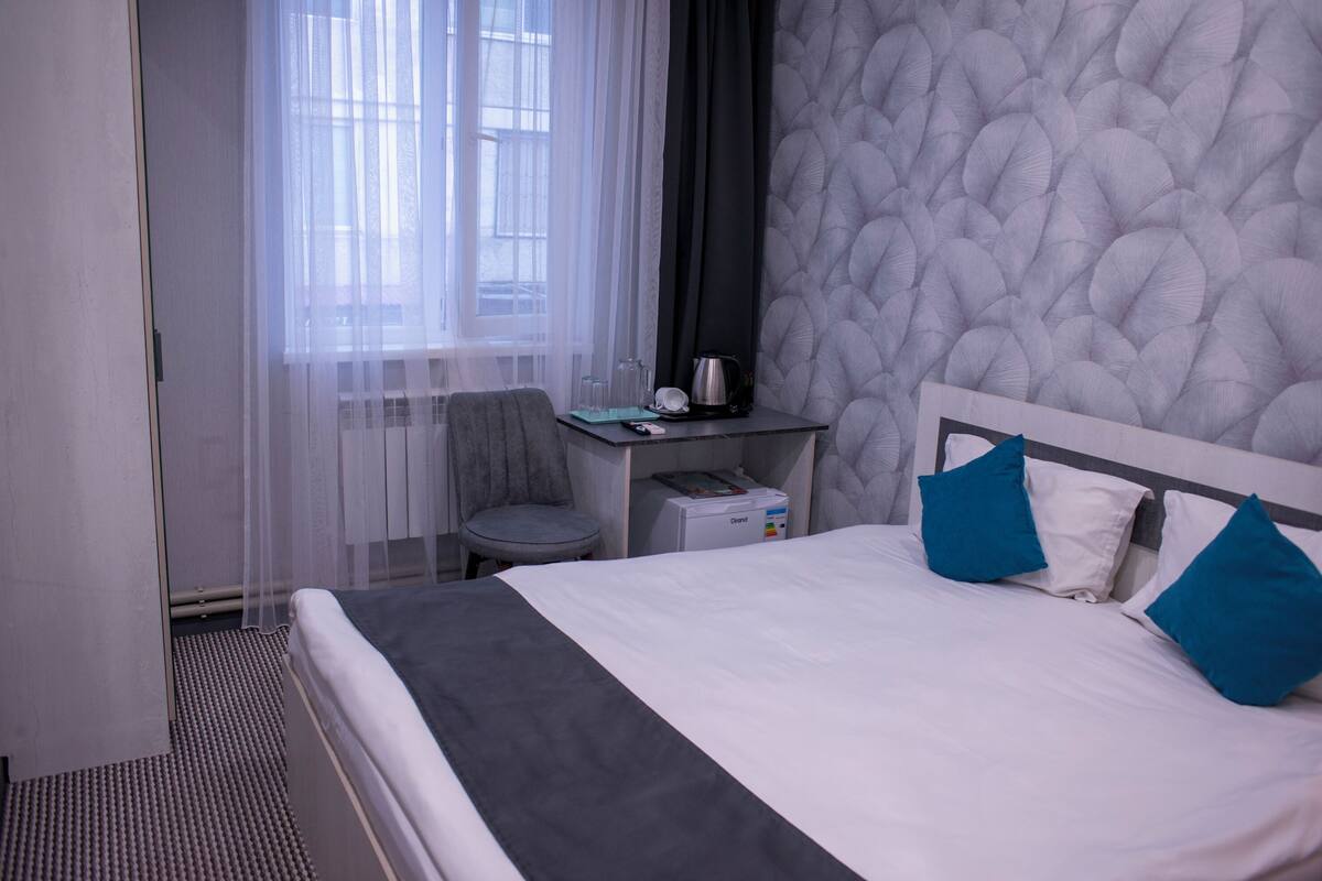 A hotel room with a bed, a desk, a chair, a window with sheer curtains, and a textured wall.