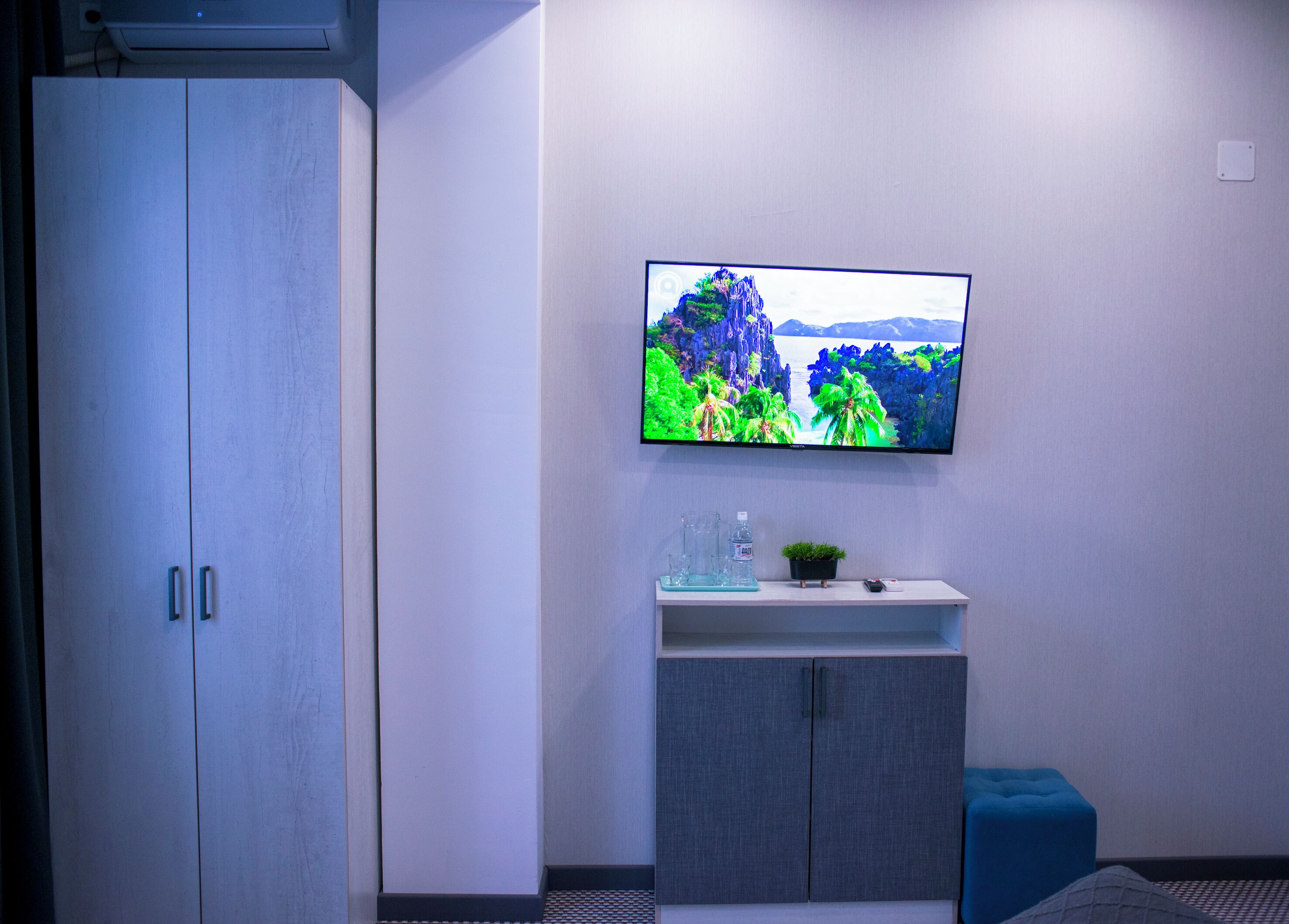 Una habitación con un televisor montado en la pared, un mueble bajo este y un otoman azul.