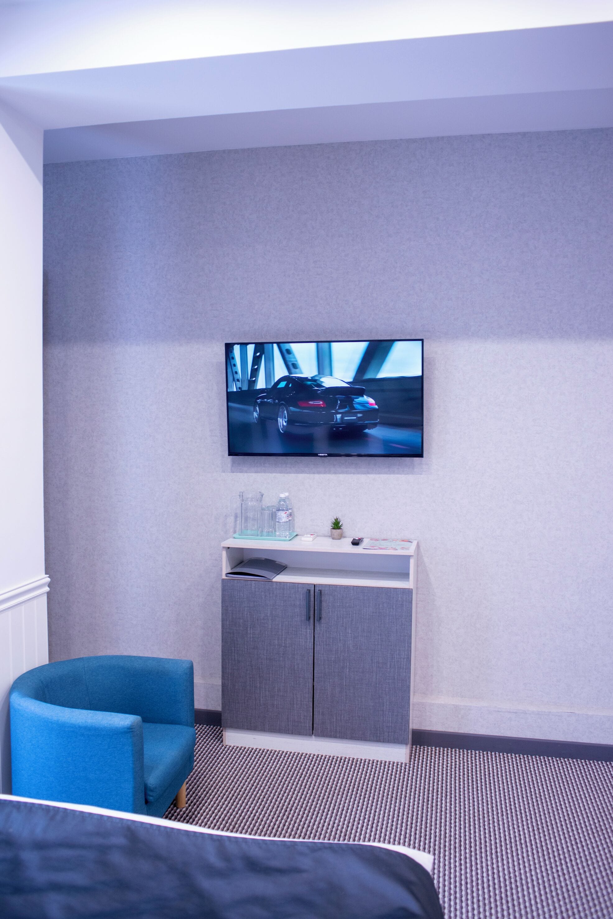 A hotel room with a bed, a blue armchair, a TV mounted on the wall, and a small desk with a plant.
