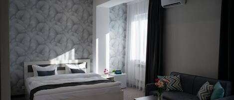 A modern hotel room with a bed, a sofa, a small table, and an air conditioning unit.