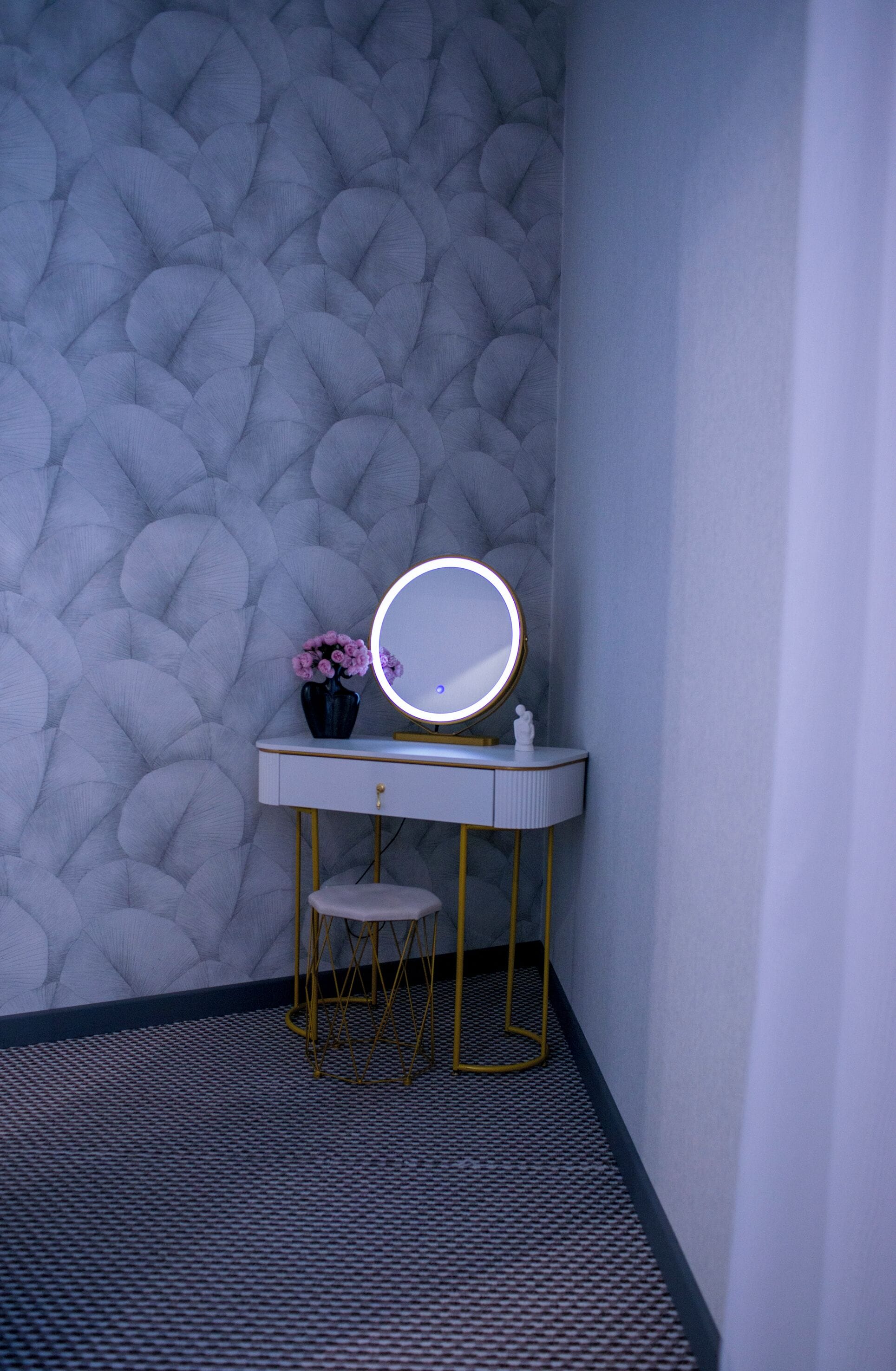 A bathroom with a textured wall, a vanity with a round mirror, a small stool, and a vase with flowers.