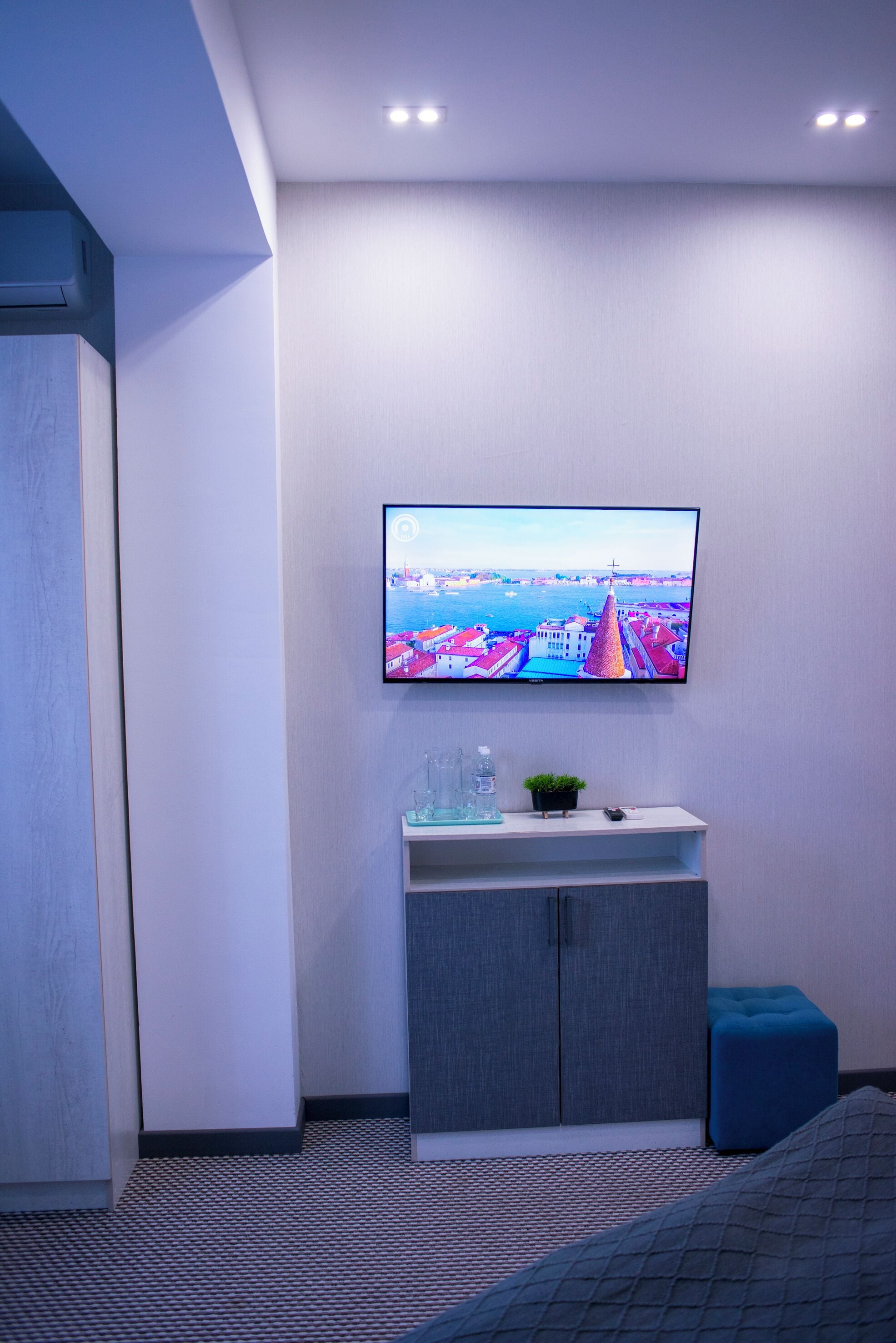 Una habitación de hotel moderna con un televisor de pantalla plana montado en la pared, un pequeño mueble con una planta y un otoman azul.