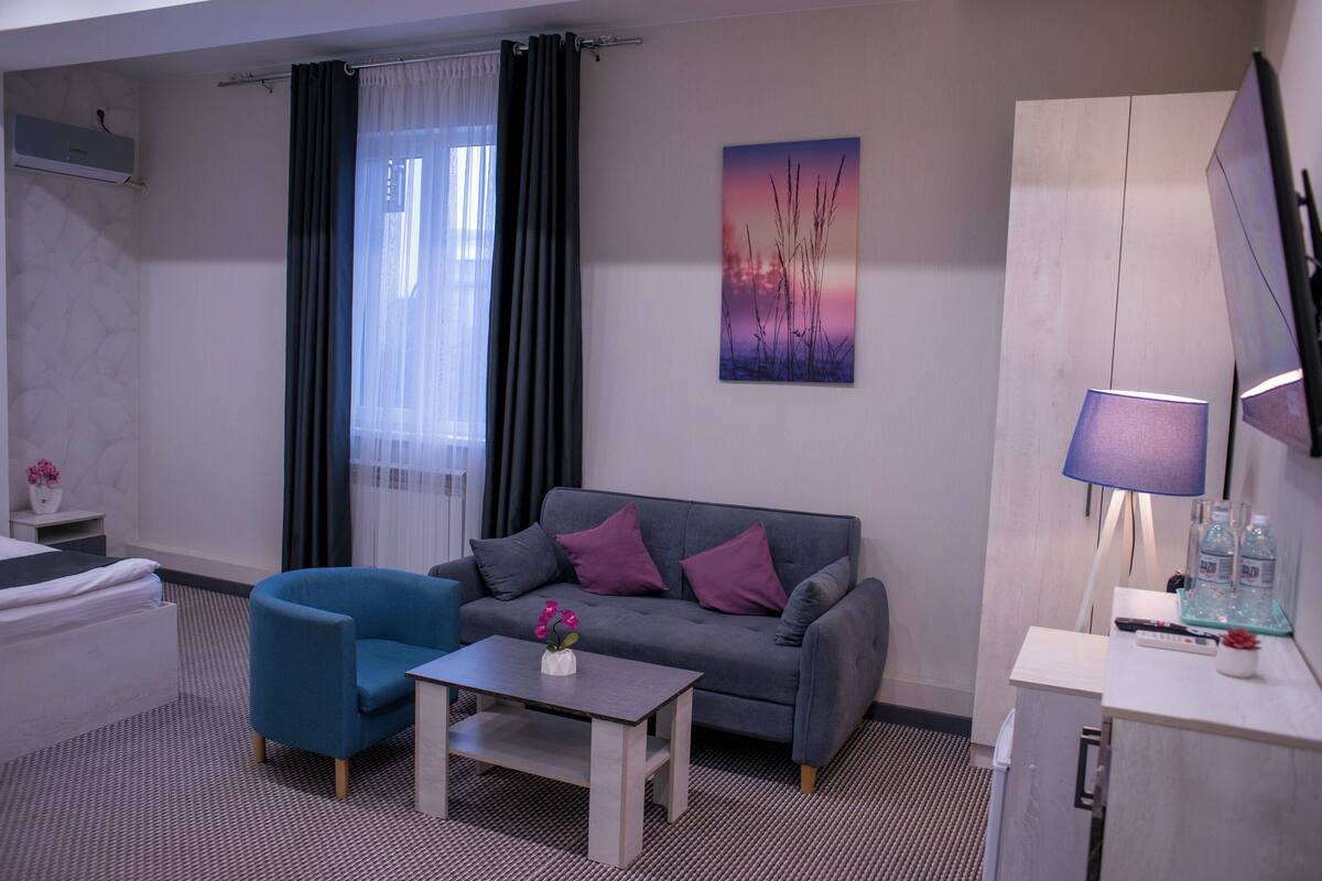 A modern hotel room with a grey sofa, a blue armchair, a small table with a vase, a TV, and a cabinet.