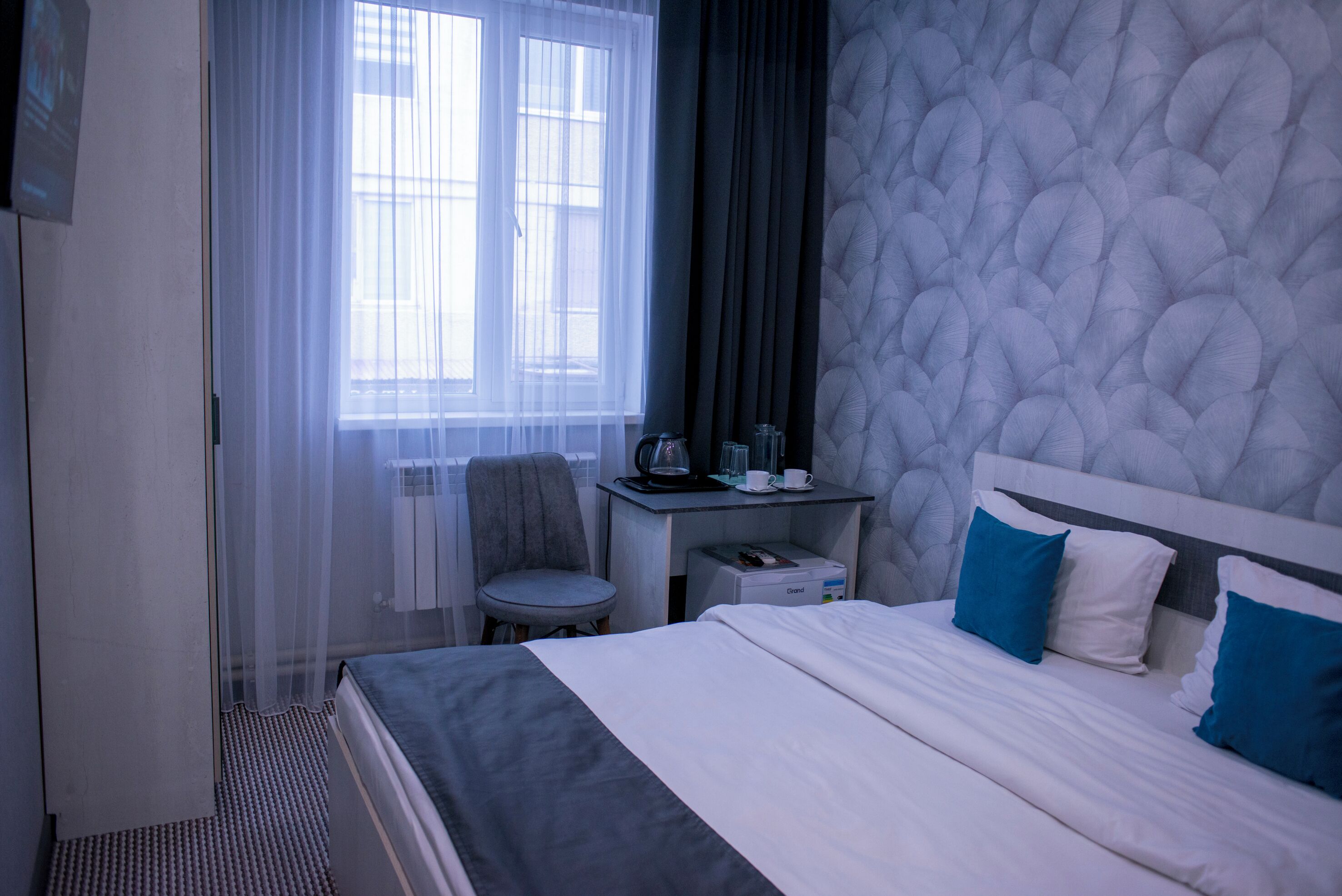 A hotel room with a bed, a desk, a chair, and a window with sheer curtains.