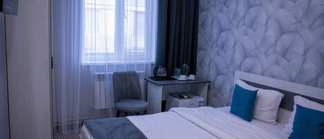 A hotel room with a bed, a desk, a chair, and a window with sheer curtains.
