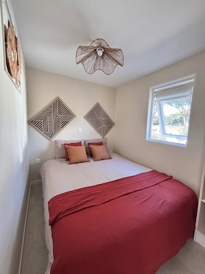 A bedroom with a bed, pillows, a red blanket, two wall-mounted decorative panels, a ceiling light fixture, and a window with a view.
