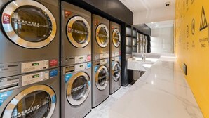 A row of stacked, stainless steel front-loading washers with control panels, in a modern hotel lobby with marble flooring and a yellow accent wall. - Atour S Hotel  Hongqiao Center Shanghai (Shanghai)